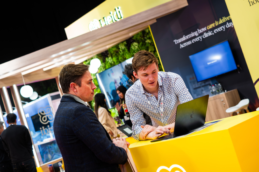 A man in a blue blazer is conversing with another man in a checkered shirt at a booth with a bright yellow counter and promotional displays in the background. The second man is working on a laptop.