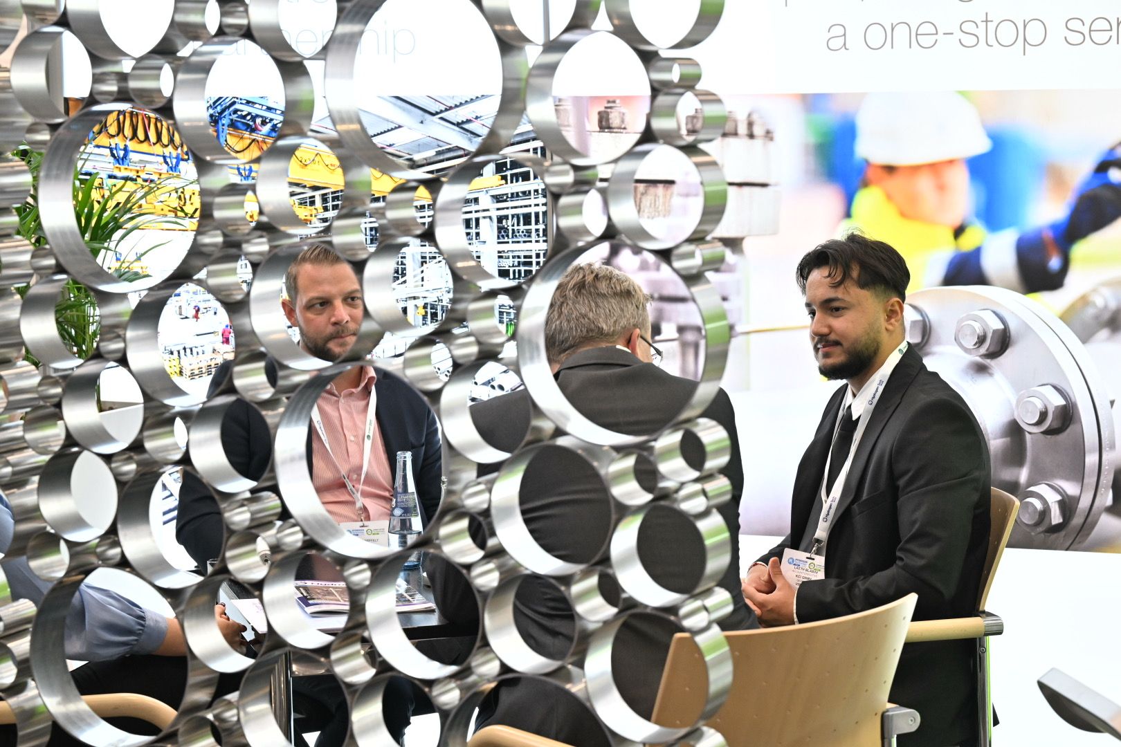 Three men in business attire have a meeting at a trade show booth, seen through a decorative metal screen with circular openings.