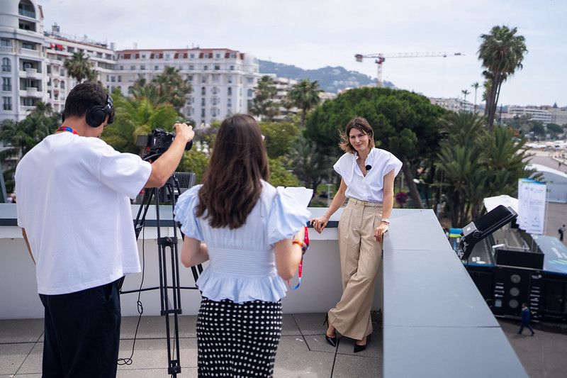 A woman in a white blouse and tan trousers stands against a rooftop railing, smiling during an outdoor interview. In the foreground, a videographer with headphones operates a camera on a tripod next to an interviewer seen from behind.