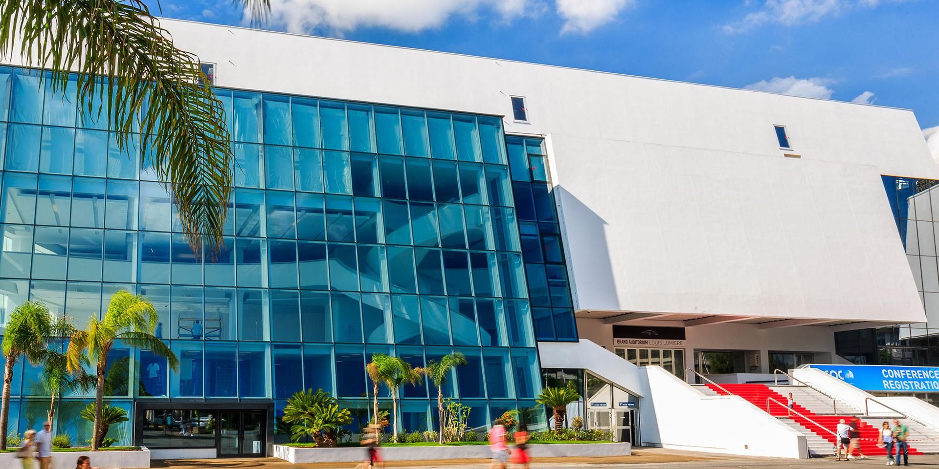 The exterior of the Palais des Festivals in Cannes, featuring a large blue glass facade, a white modern building structure, palm trees, and a red carpet on the entrance stairs.
