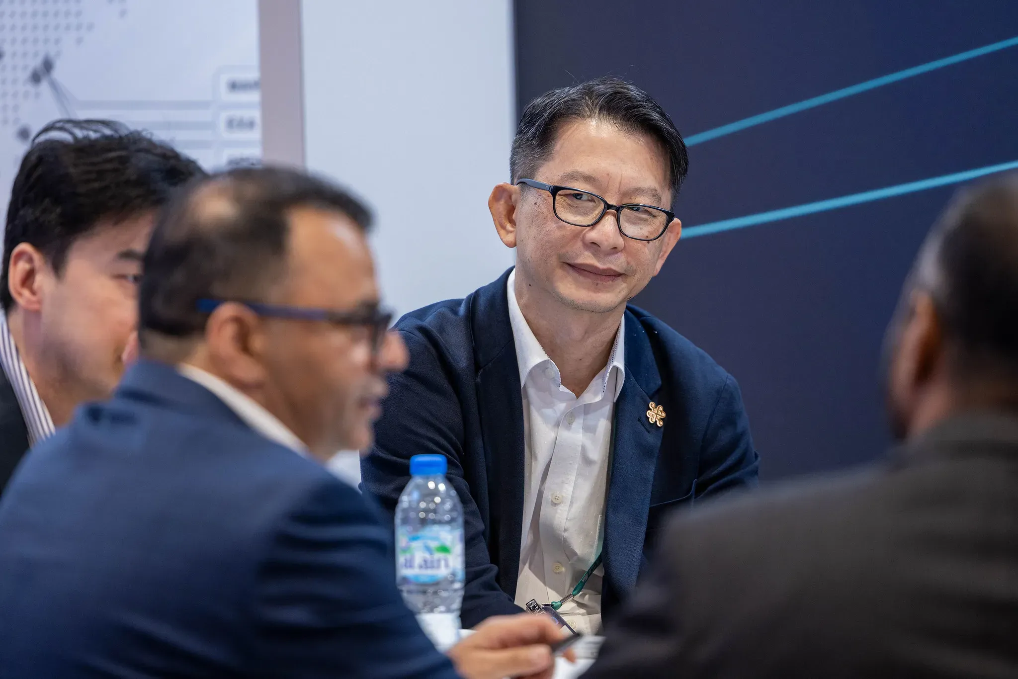 An Asian businessman wearing black-rimmed glasses and a dark blue blazer looks on with a calm smile during a group discussion around a conference table.