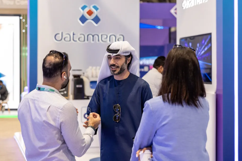 An Emirati businessman in a blue thobe and white ghutra smiles while networking with two attendees at an exhibition booth featuring the 