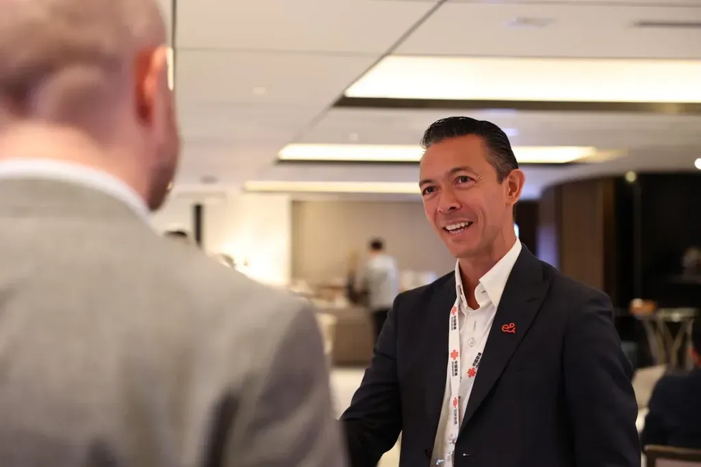 A professional man in a dark suit jacket and white dress shirt smiles while conversing with a colleague whose back is to the camera. He wears an event lanyard and a small red lapel pin.