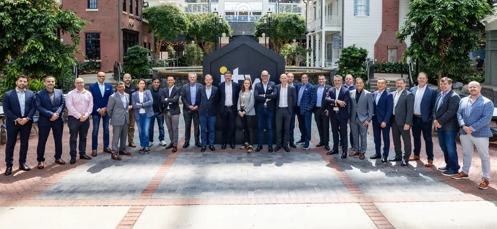 A large group of business professionals standing in a wide line for a group photograph outdoors at ITW 2025. They are posed in front of a large, dark house-shaped sign featuring the 