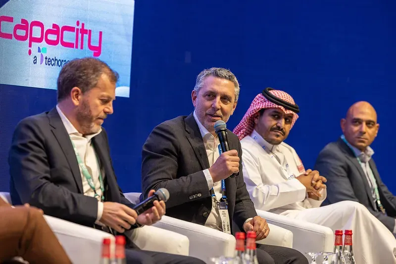 Four male executives participate in a panel discussion on a stage. The panelist in the center speaks into a microphone while his colleagues listen. Part of the magenta 