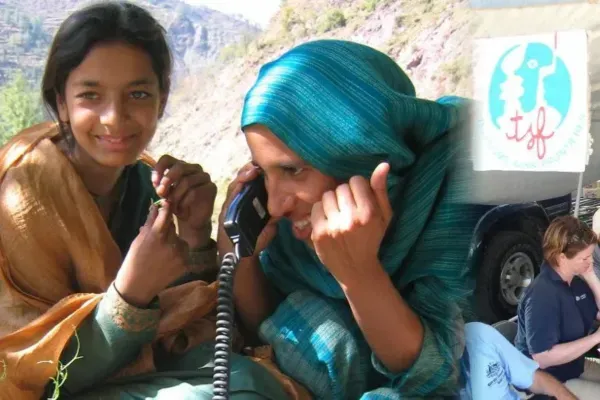A composite image showing a young girl smiling next to a woman in a teal headscarf who is joyfully talking on a satellite phone handset outdoors in a mountainous region. The right side features a T&eacute;l&eacute;coms Sans Fronti&egrave;res (TSF) banner and field workers.