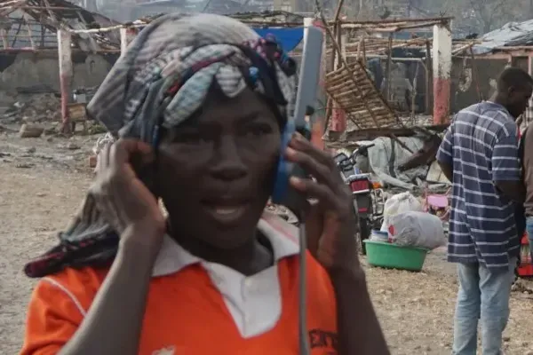 A person wearing an orange shirt and a patterned headwrap speaks on a satellite phone handset, standing outdoors in front of an area with heavily damaged structures and debris.