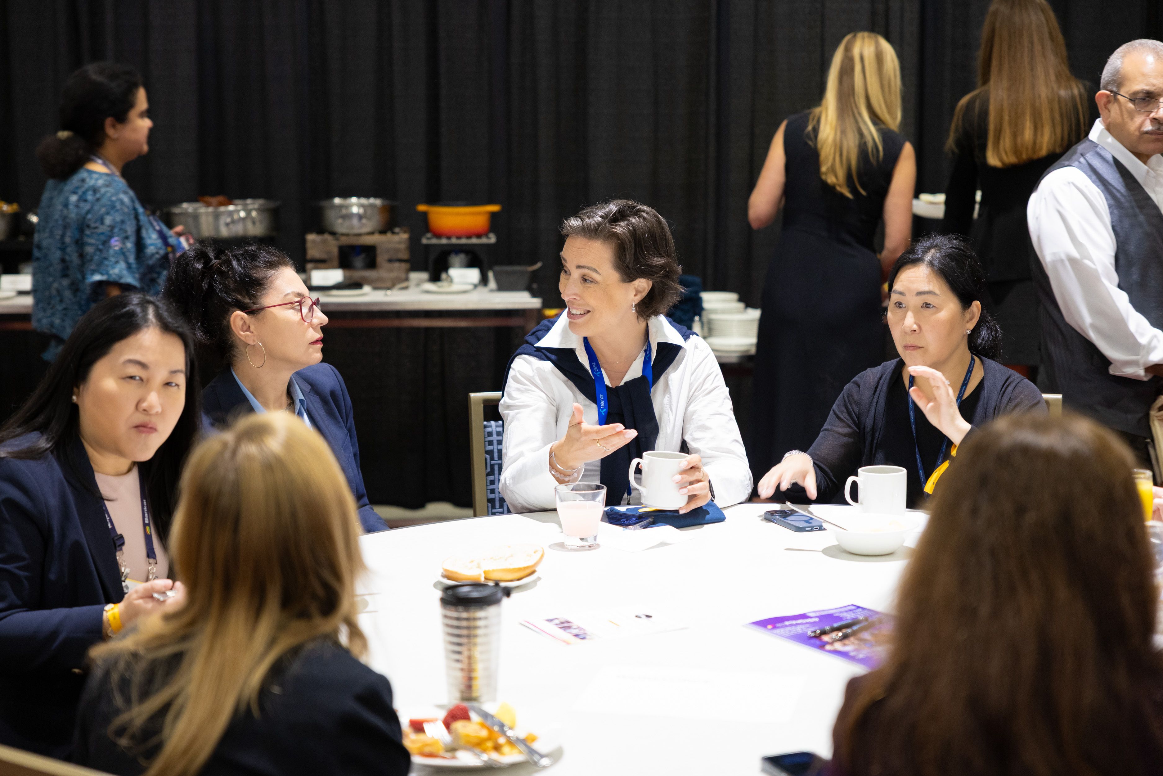 A group of professional women sit around a white circular table, engaged in a lively discussion during a breakfast meeting. They hold coffee mugs amidst plates of fruit and pastries.