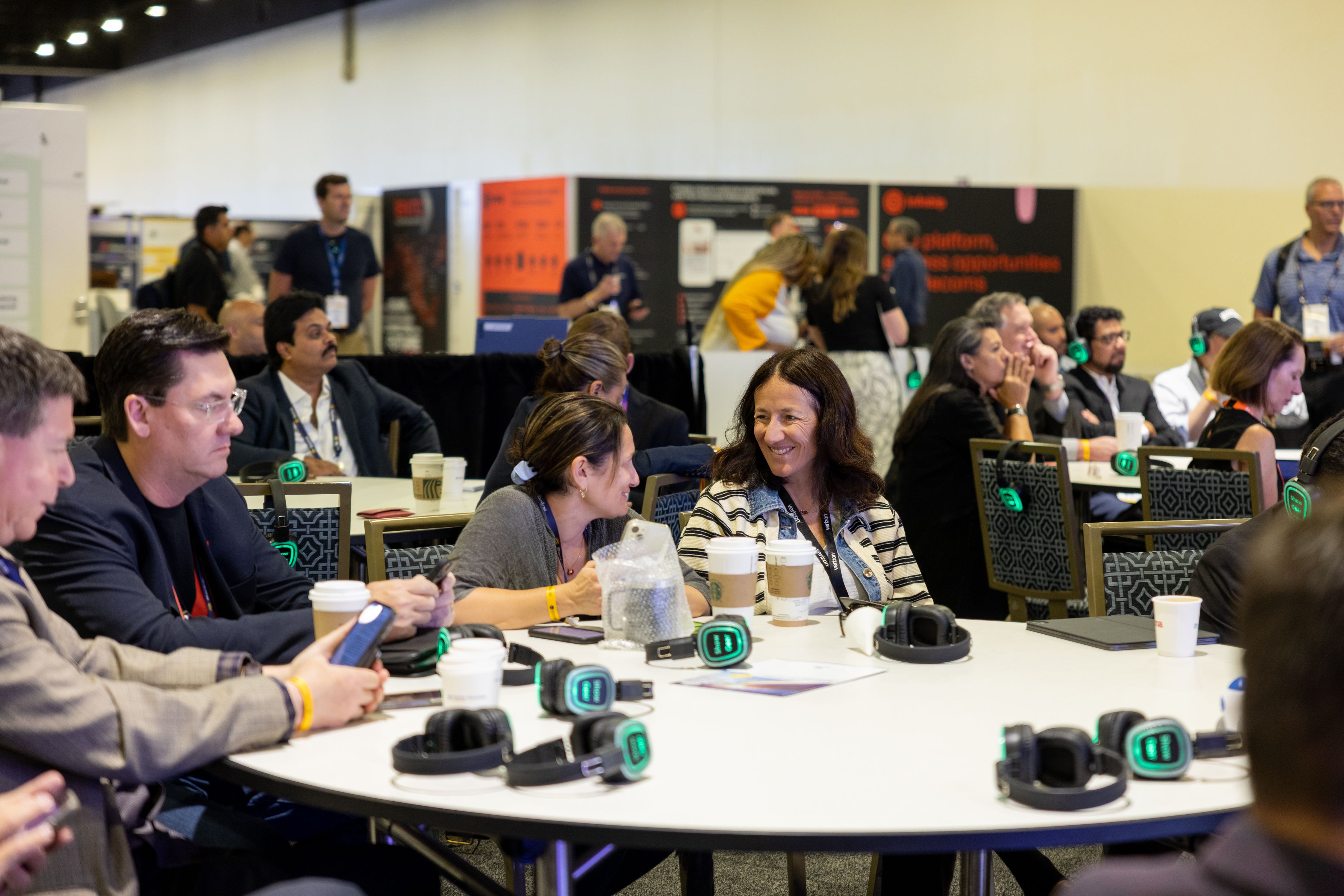 Attendees sit at round tables in a large conference hall, some wearing luminous green headphones. Coffee cups and headsets are scattered across tables during a collaborative networking or listening session.