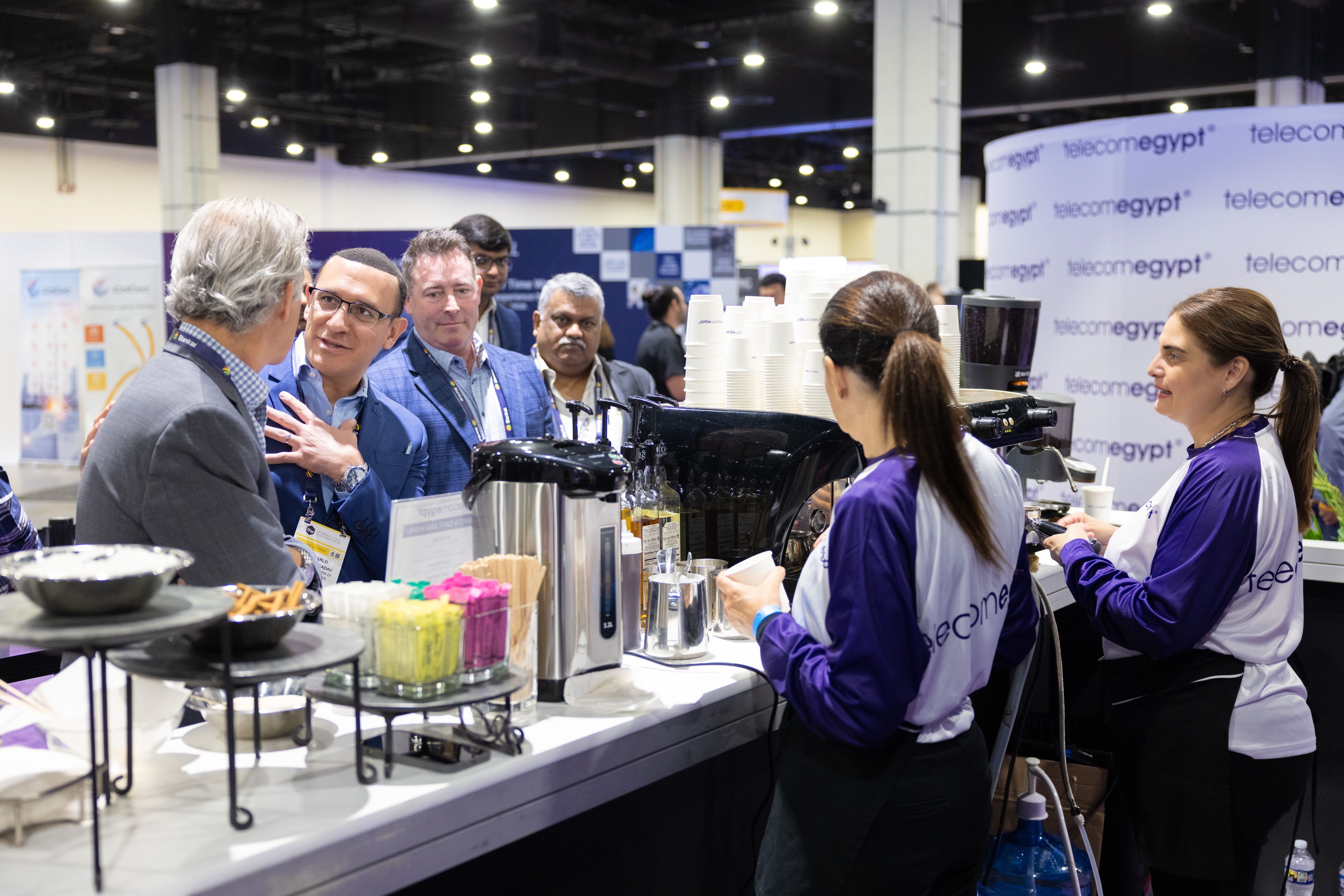 A busy Telecom Egypt booth at a conference features staff serving coffee from a white counter to several men in business attire, set against a branded, professional exhibition hall background.