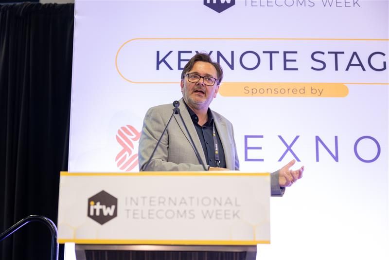 A man in a grey blazer and glasses gestures while speaking at a podium labeled "ITW International Telecoms Week. He stands on a stage with a backdrop.