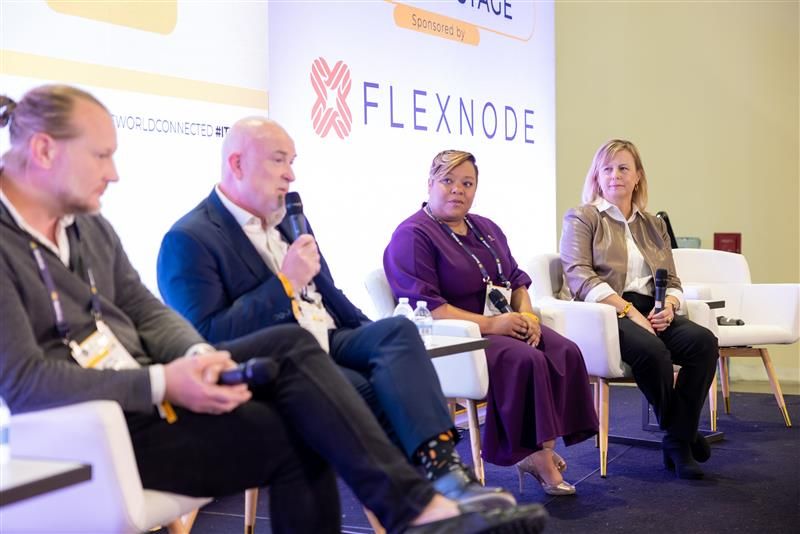 Four speakers participate in a professional panel discussion on stage. They sit in white chairs before a Flexnode backdrop, with one man speaking into a microphone while others listen attentively.