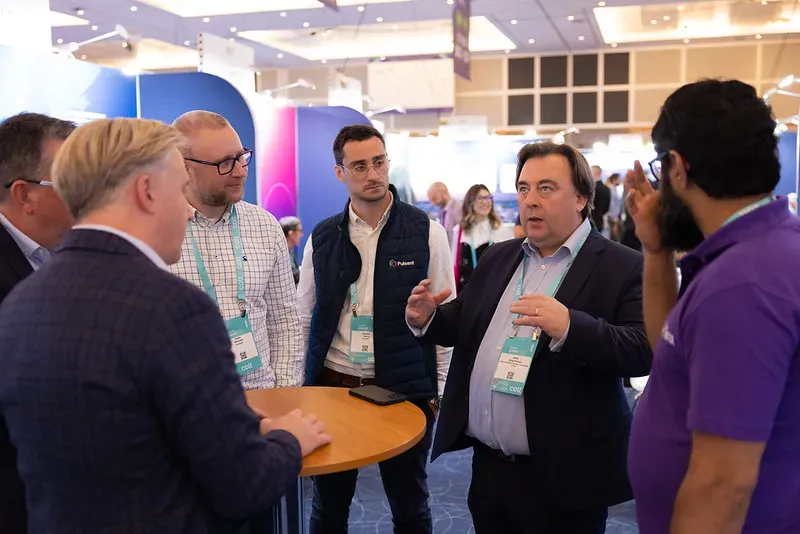 A group of professionals in business attire engaged in an active discussion around a high-top table at the Capacity Europe conference exhibition hall.