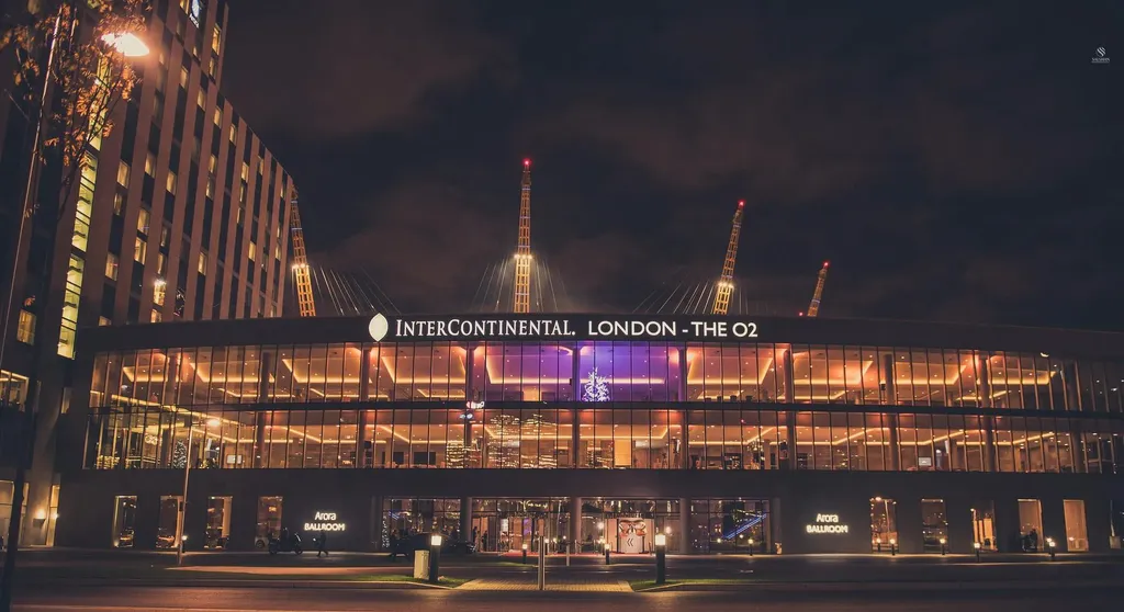 A wide night-time shot of the InterContinental London – The O2. The modern glass-fronted building is brightly lit with purple and warm interior lights, with the illuminated white signage 