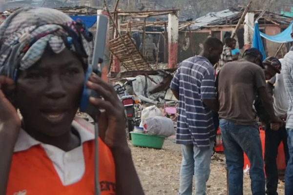 A person in a bright orange shirt and a headwrap uses a large satellite phone in the foreground of a damaged area. In the background, other people stand amidst debris and makeshift structures, illustrating T&eacute;l&eacute;coms Sans Fronti&egrave;res' work in emergency response zones.