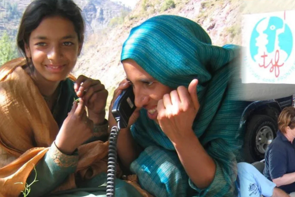 A smiling girl looks at the camera while a woman next to her, wearing a teal headscarf, smiles and speaks into a corded satellite phone. In the background, the T&eacute;l&eacute;coms Sans Fronti&egrave;res (TSF) logo is visible on a white banner, signifying the organization's role in providing vital communication links.