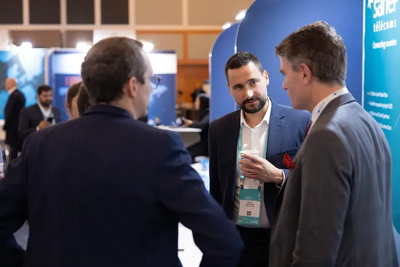 Three business professionals in suits networking at a Capacity Europe event. They are standing near exhibition booths, wearing event lanyards and engaged in conversation in a brightly lit conference hall.