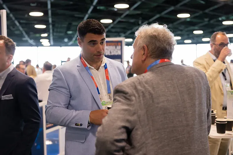 A photograph from a professional networking event showing a man in a light blue blazer and white dress shirt engaged in a focused conversation with another man whose back is to the camera. They are standing in a large, brightly lit convention hall with other attendees, networking tables with coffee cups, and event signage visible in the blurred background. Both men are wearing event lanyards, capturing a candid moment of business interaction at Capacity Europe.