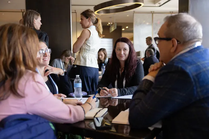 A group of business professionals sitting around a conference table engaged in a meeting, with other attendees mingling in the background of an event lounge.