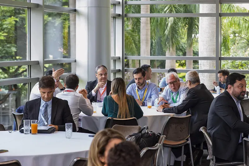 A group of professionals in business attire conversing around large circular tables at a conference luncheons or networking event, set against large glass windows looking out at palm trees.
