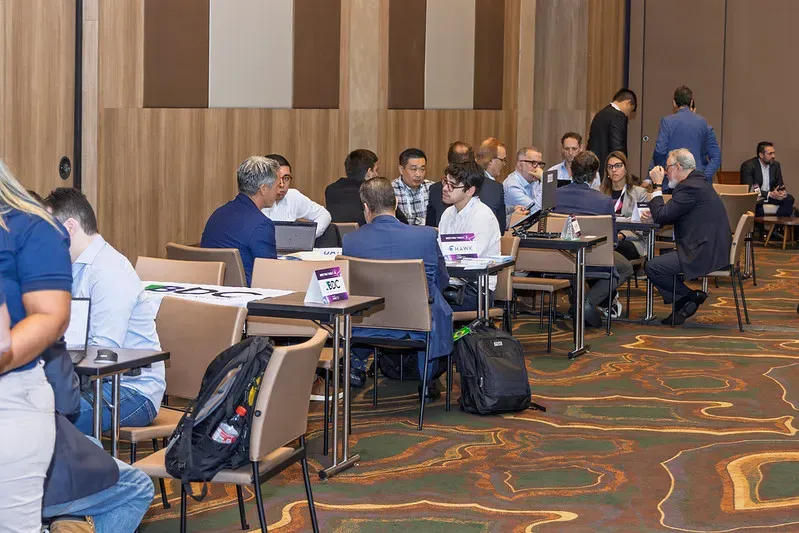 A large conference room filled with professionals seated at small tables, engaged in business meetings and networking discussions.