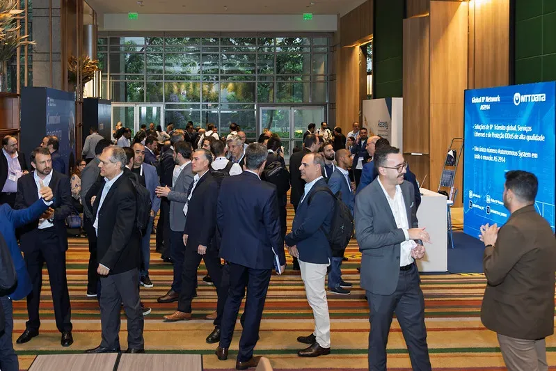 A crowded networking floor at a technology conference, showing professionals in business attire mingling near sponsor booths, including a bright blue NTT DATA display banner.