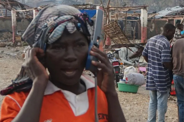 A close-up photograph of a person wearing an orange shirt and a patterned head covering, holding a large grey satellite phone with a prominent antenna to their ear. The individual has an intense expression while communicating. The background shows a scene of severe destruction, with collapsed wooden structures, debris-strewn ground, and a green plastic basin. In the background, other individuals stand among the remnants of the damaged settlement.