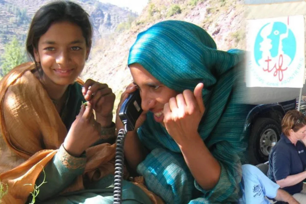 A composite photograph capturing a humanitarian aid scene in a rural, mountainous landscape. On the left, a young woman with dark hair and a gold shawl smiles warmly at the camera. Next to her, a woman wearing a teal headscarf smiles joyfully while speaking into a black satellite phone handset with a coiled cord.  The right side of the image features a blended overlay showing a white banner with the blue and red logo of 