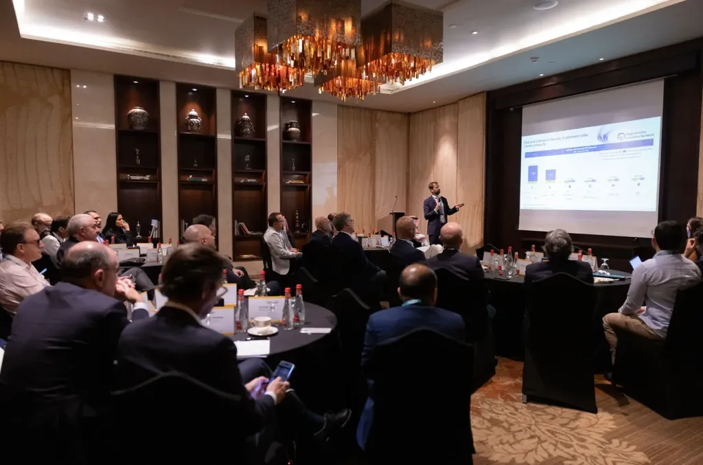 A speaker stands at the front of a conference room presenting a slideshow to an audience of digital infrastructure executives seated at round tables during the MENA Digital Infra Leaders Summit.