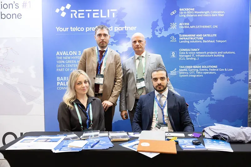 Four corporate professionals posing together at an exhibition stand for Retelit. Two are seated at a black desk with brochures, and two stand behind them in front of a blue banner displaying company services.