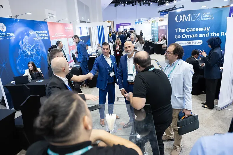A high-angle shot of an exhibition aisle at a busy trade show. In the center, two businessmen in bright blue suits shake hands and network with other attendees near an OMIX booth.