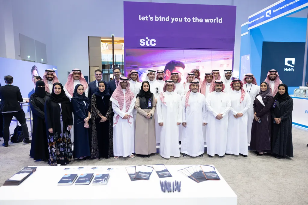 A wide group portrait of corporate professionals standing together in front of a purple exhibition booth branded with the 