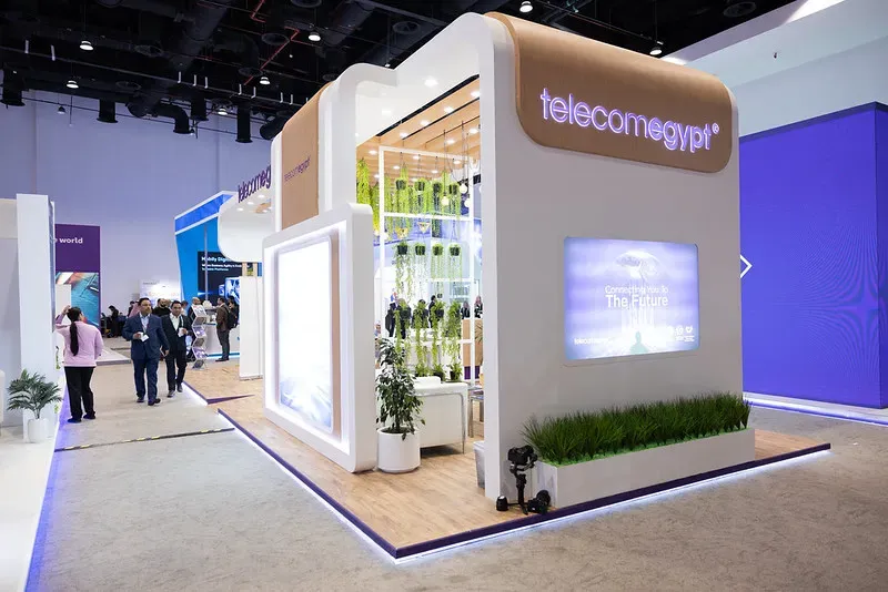 A large, modern, custom-built multi-level exhibition booth for Telecom Egypt at a convention center, featuring wood-textured flooring, white walls, built-in digital screens, and hanging green plants.