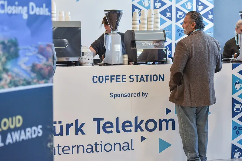 A man stands waiting at an event coffee station sponsored by T&uuml;rk Telekom International. The station features professional espresso machines, coffee grinders, and stacked paper cups.