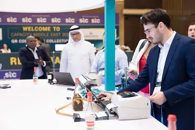 An attendee in a dark blue blazer interacts with a desktop badge printer at a registration desk during Capacity Middle East, with stc-branded welcome signage visible in the background.