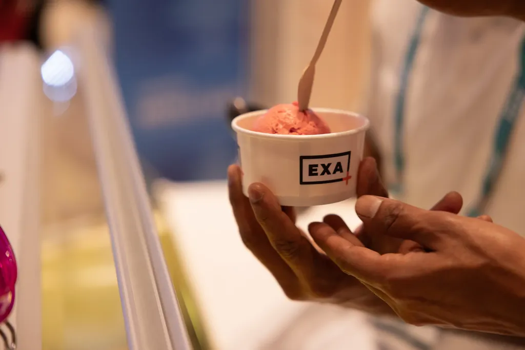 A close-up shot of a person's hands holding a white paper cup containing a scoop of pink ice cream or sorbet. The cup prominently features a black rectangular logo that reads 