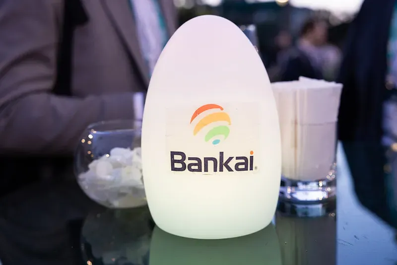 A close-up shot of an egg-shaped, glowing white table light sitting on a dark glass table during an evening event. The lamp features a crisp corporate logo for 