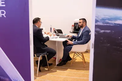 A view through a doorway into a private, white-walled meeting space. Two businessmen in suits are seated opposite each other at a small white table, deep in conversation with a laptop and documents open between them.