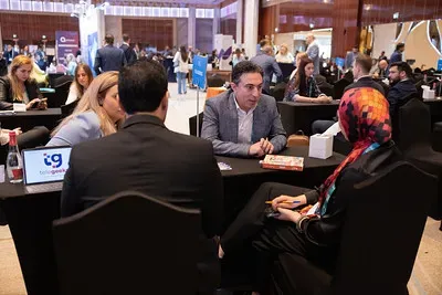 An indoor view of a busy conference hall arranged with rows of black-clothed meeting tables. In the center, several professionals are engaged in face-to-face discussions, including a man in a grey patterned blazer speaking to a woman wearing a vibrant, geometric-patterned hijab.