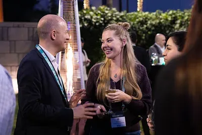 An outdoor evening networking reception where a man and a woman stand close to a tall, flaming pyramid patio heater, smiling and chatting. The woman holds a glass of white wine and wears an event lanyard. Green hedges and the silhouettes of other attendees are visible in the background.