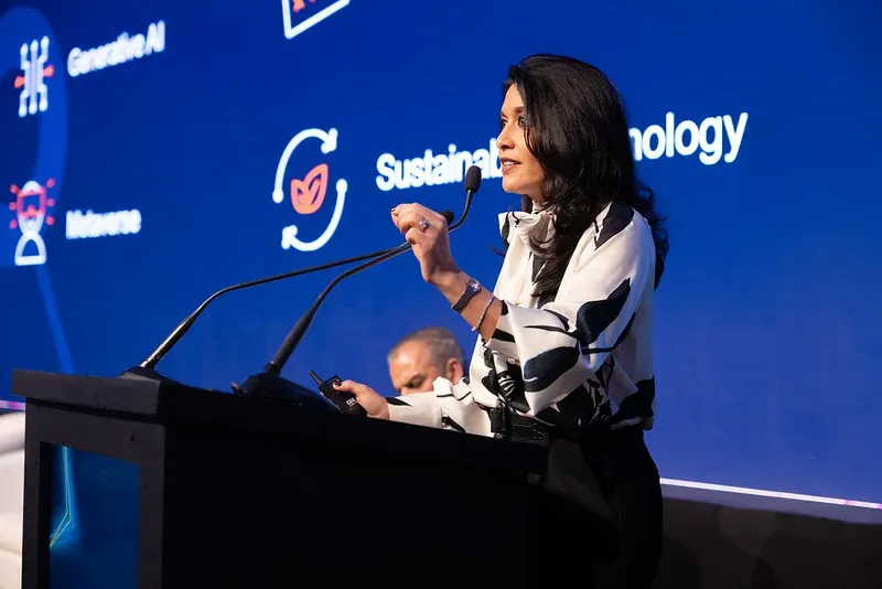 A professional woman with dark hair stands at a black podium, speaking into a microphone on a stage. She is wearing a white blouse with a bold black abstract pattern. Behind her is a large blue digital screen displaying corporate icons and white text, including the words 