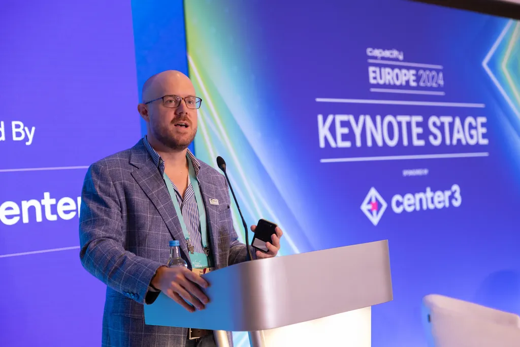 A speaker stands at a modern, metallic podium on a stage. The man is bald, has glasses and a beard, and is wearing a blue-and-white patterned blazer over an open-collar striped shirt with an event lanyard. He looks toward the audience while holding a smartphone in his left hand. Behind him is a large blue digital screen displaying text that reads 