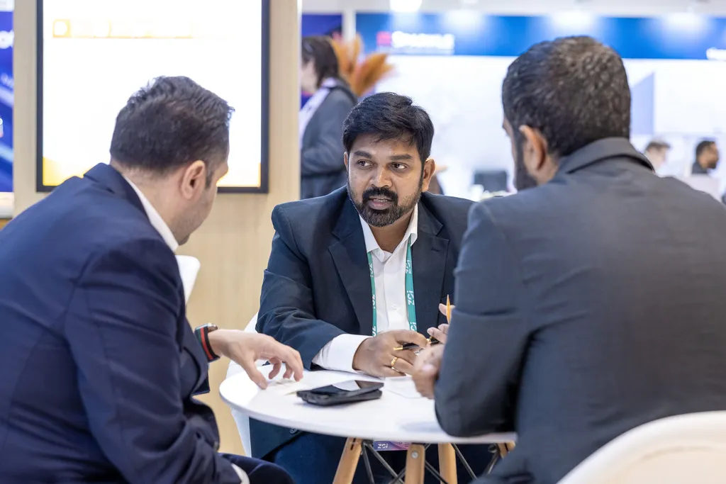 A candid shot of three professionals conducting a meeting around a small white table at a convention. The central businessman with a neat beard and a dark suit looks toward his colleague while speaking and holding a pen.