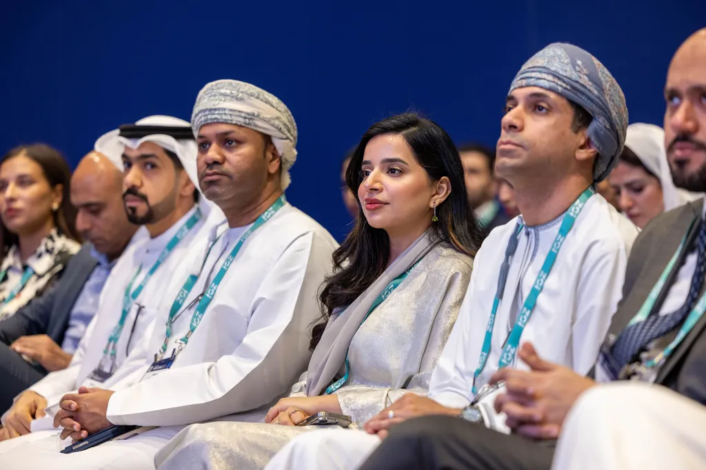 A side-angle shot of an attentive audience seated in rows at a conference. The group includes Middle Eastern professionals in formal attire, including men wearing traditional Omani muzzar turbans and a woman in a shimmering grey abaya, all looking toward the stage.
