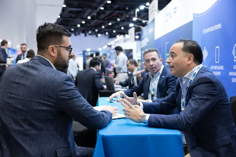 Two businessmen seated across from each other at a blue-clothed table, engaged in an active discussion during a busy corporate exhibition. The man on the right gestures with his hands as he speaks.