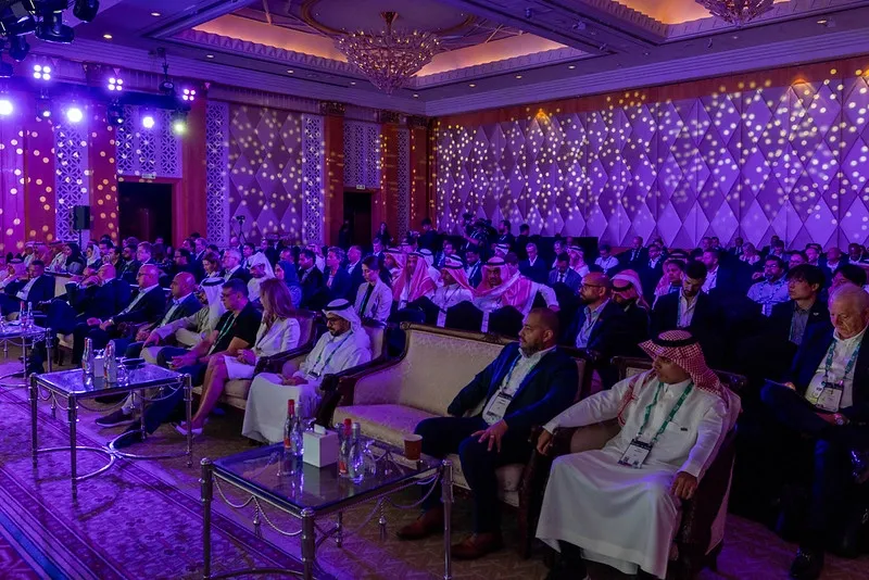 A large audience is seated in a grand, dimly lit ballroom illuminated by purple lighting and dotted light projections on the walls. Several chandeliers hang from the ornate ceiling. In the foreground, people are seated on couches and chairs facing forward, with low glass tables in front of them