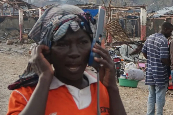 A person wearing an orange shirt and a patterned headwrap speaks on a satellite phone handset, standing outdoors in front of an area with heavily damaged structures and debris.