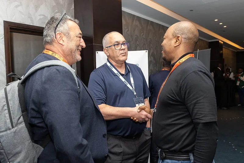 Three gentlemen standing. One is shaking hands and he's wearing a lanyard, one is smiling. 