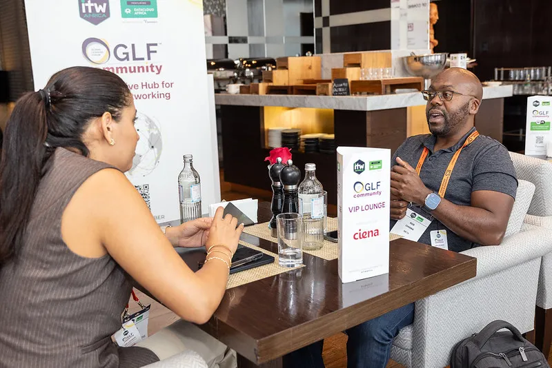 Two people sitting on a comfy chair, discussing a topic. Behind them is a food and drink station, on the table there is a GLF sign and a banner on the left. 