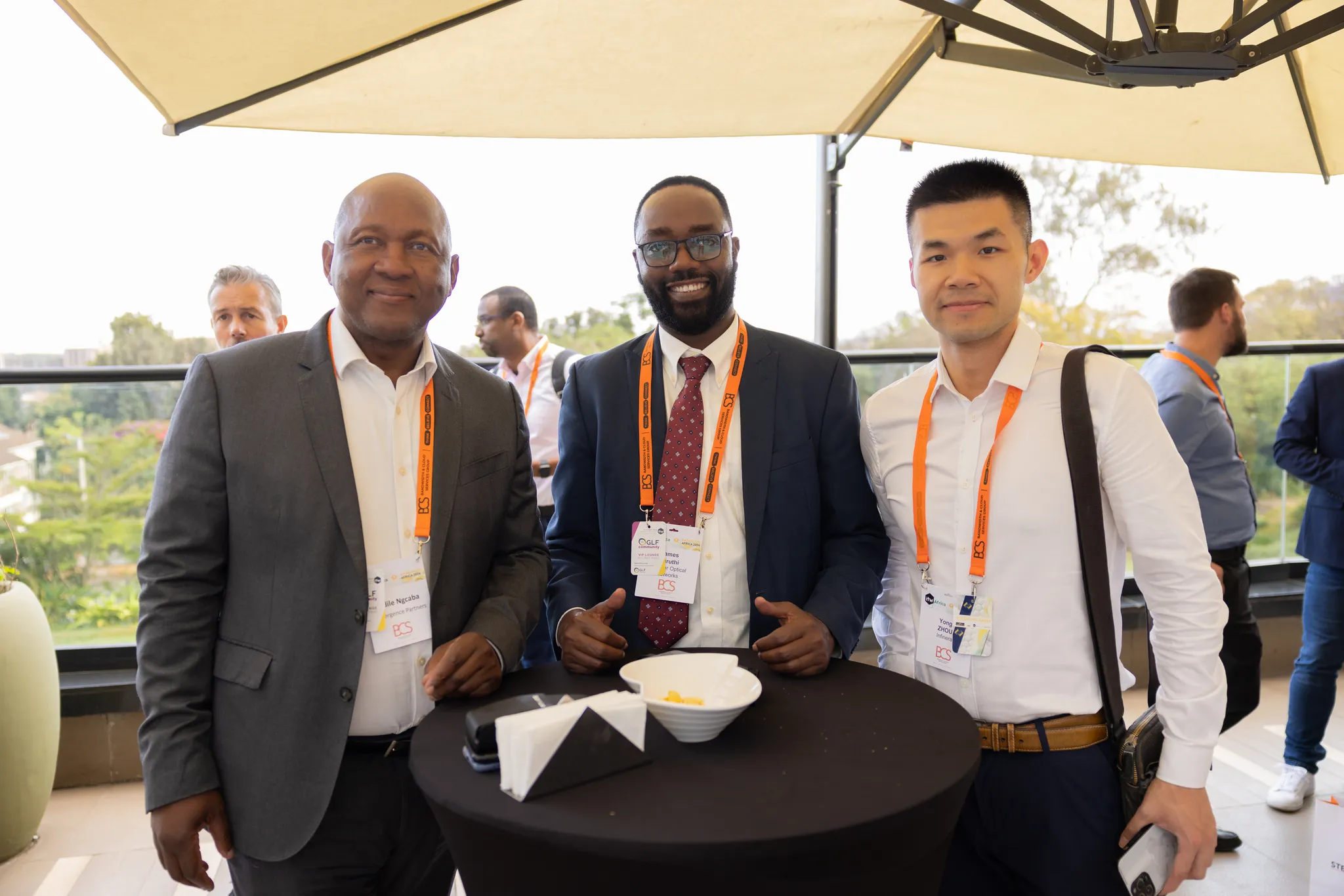 Three attendees in business attire wearing event lanyards smiling and posing for a photo around a cocktail table at an outdoor networking reception.